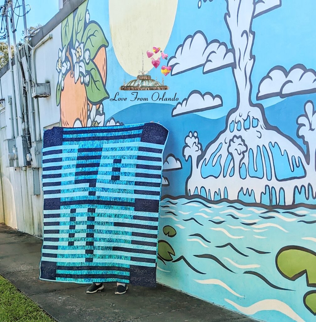 Me holding up the blue Through Love Quilt in front of an Orlando mural.