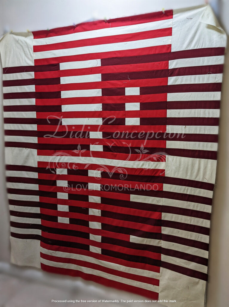 Through Love Quilt top in deep red, medium red and cream, hung up on my design wall.